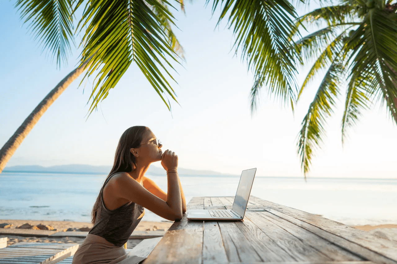 A remote worker pauses beside a laptop on a tropical beach, framed by palm trees and ocean.